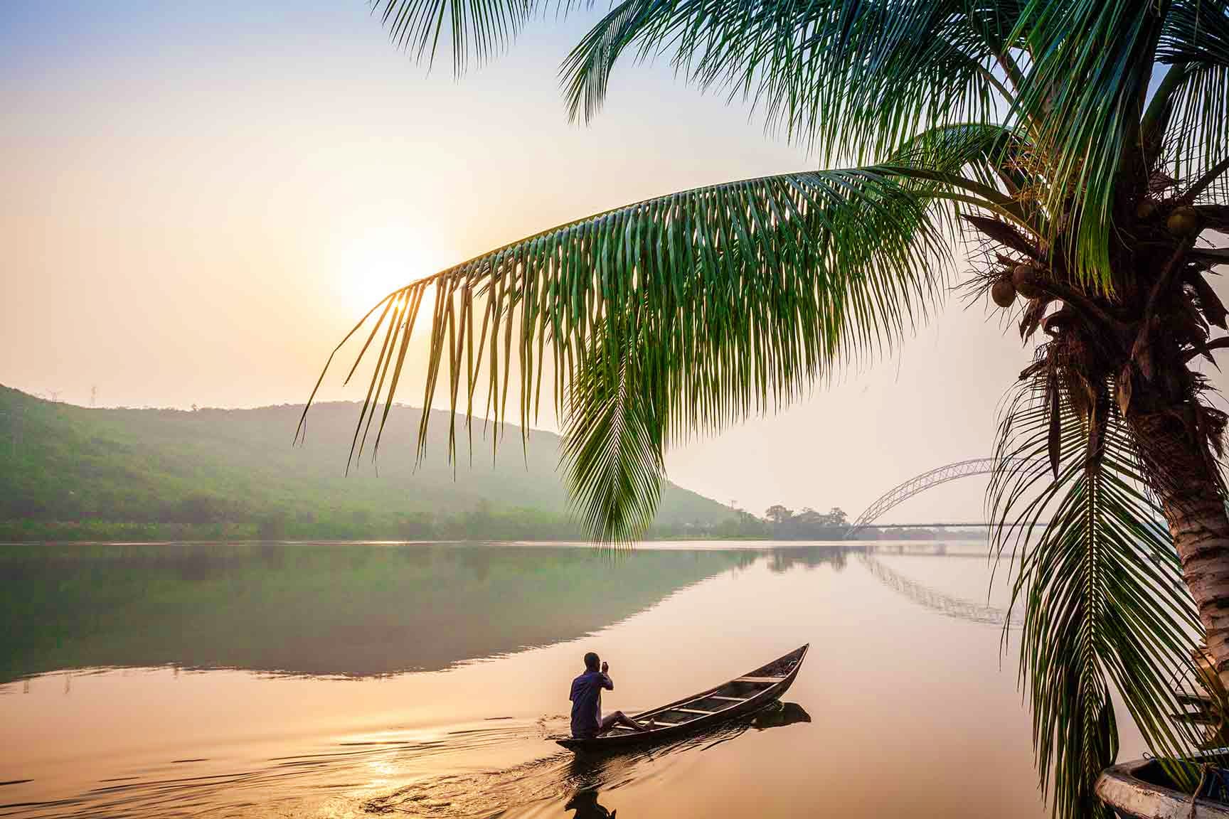 Person in boat on calm river at sunrise with palm fronds and bridge in background, Ghana landscape